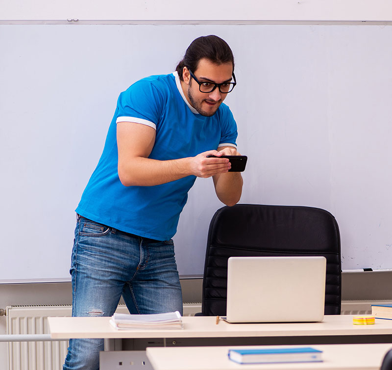 Un homme dans une salle de classe prenant des photos avec un smartphone d'un ordinateur portable.